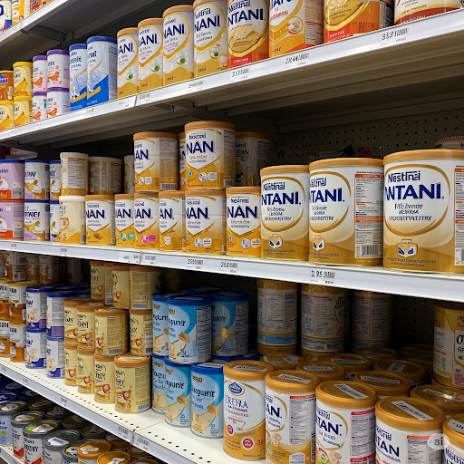 Prateleira de supermercado repleta de diversas latas de leite em pó infantil, incluindo marcas populares como Aptamil, NAN, Milnutri, Nestogeno e Ninho. Esta seção destaca o foco Melhores produtos para Bebê na nutrição infantil.